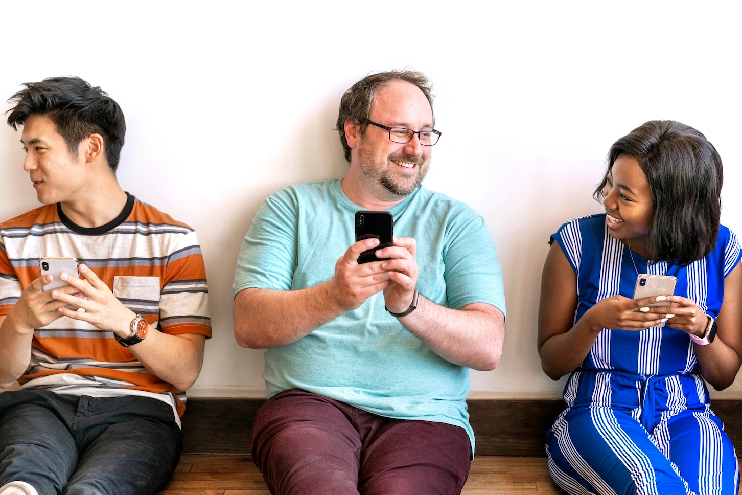 Three people sat against a wall, smiling and looking at phones